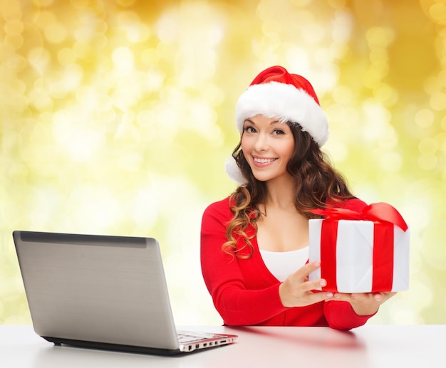 concepto de navidad, vacaciones, tecnología y personas: mujer sonriente con sombrero de ayudante de santa con caja de regalo y computadora portátil sobre fondo de nieve amarillo