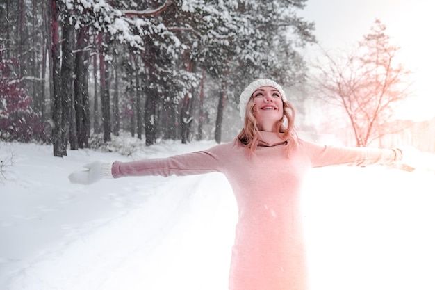 Concepto de Navidad, vacaciones y temporada. Joven mujer feliz que sopla nieve en la naturaleza del bosque de invierno. Ropa de abrigo guantes de punto y gorro. Fondo de paisaje de bosque de invierno