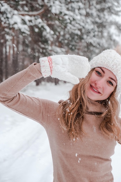 Concepto de Navidad, vacaciones y temporada. Joven mujer feliz que sopla nieve en la naturaleza del bosque de invierno. Ropa de abrigo guantes de punto y gorro. Fondo de paisaje de bosque de invierno