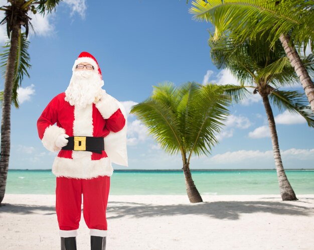 concepto de navidad, vacaciones, viajes y personas - hombre disfrazado de santa claus con bolsa sobre fondo de playa tropical