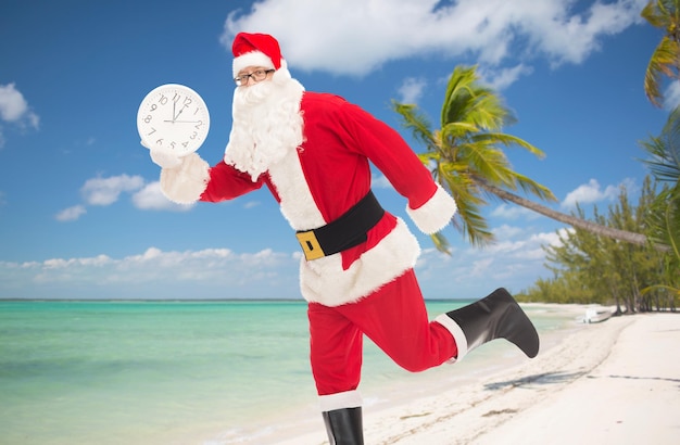 concepto de navidad, vacaciones, viajes y personas - hombre disfrazado de santa claus corriendo con un reloj que muestra doce sobre fondo de playa tropical