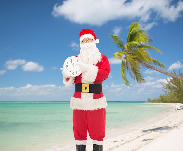 concepto de navidad, vacaciones, viajes y personas - hombre disfrazado de santa claus con reloj mostrando doce dedos apuntando sobre fondo de playa tropical