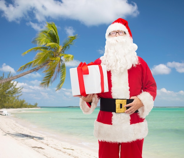 concepto de navidad, viajes, vacaciones y personas - hombre disfrazado de santa claus con caja de regalo sobre fondo de playa tropical