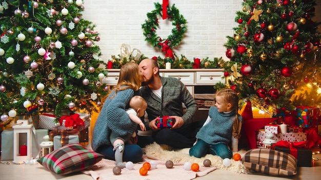 Concepto navideño familia feliz se sienta en el entorno navideño en la sala de estar e intercambian regalos mamá y papá besándose