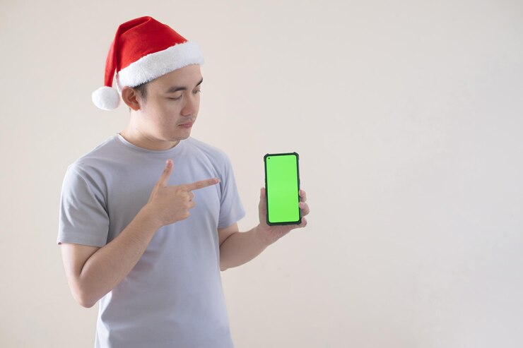 El concepto navideño de un joven asiático está apuntando a un teléfono inteligente de pantalla verde en un fondo aislado