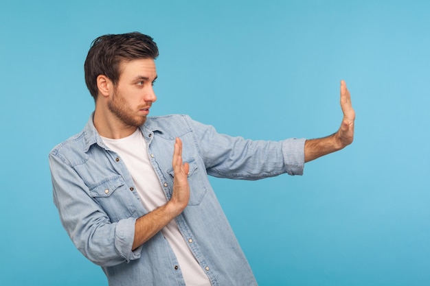 Concepto de negación de rechazo vista lateral del hombre en camisa de mezclilla de trabajador haciendo señal de alto con las manos extendidas expresión de advertencia con gesto negativo tiro de estudio interior aislado sobre fondo azul