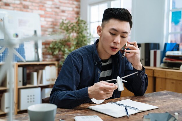 Concepto de negociación. arquitecto de hombre guapo sosteniendo el modelo de molino de viento en las manos y hablando por teléfono móvil con el cliente. trabajador tipo discutiendo el proyecto de construcción ecológica en línea en el teléfono celular en la oficina moderna.