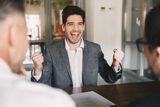 Concepto de negocio, carrera y colocación: exitoso hombre caucásico de 30 años regocijándose y apretando los puños durante la entrevista de trabajo en la oficina, con empleados de una gran empresa