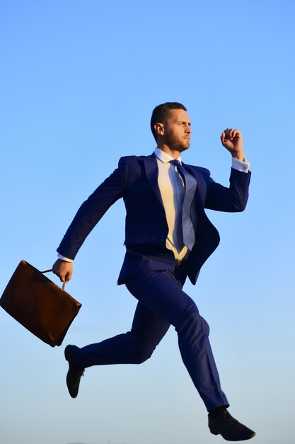 Concepto de negocio y éxito. Empresario da un gran paso en el aire. El hombre con traje formal azul sostiene una caja marrón y corre sobre un fondo de cielo azul. CEO con expresión de cara concentrada se precipita hacia adelante