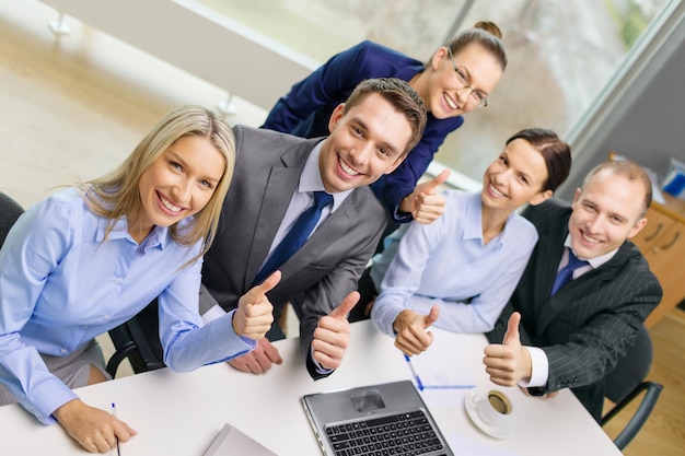 Concepto de negocio, éxito, tecnología y oficina: equipo de negocios sonriente con computadora portátil, papeles y café mostrando los pulgares para arriba en la oficina