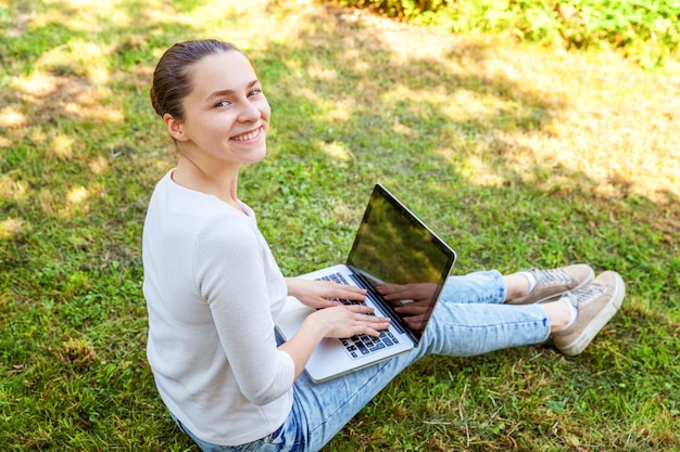 Concepto de negocio independiente. Mujer joven sentada en el césped verde en el parque de la ciudad trabajando en una computadora portátil. Estilo de vida auténtica estudiante sincera estudiando al aire libre. Oficina Móvil