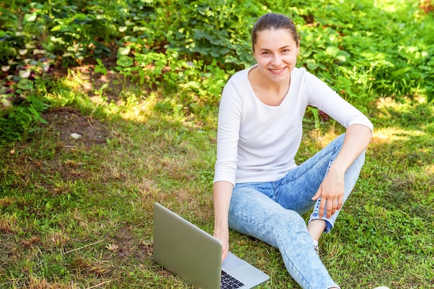 Concepto de negocio independiente. Mujer joven sentada en el césped verde en el parque de la ciudad trabajando en una computadora portátil. Estilo de vida auténtica estudiante sincera estudiando al aire libre. Oficina Móvil