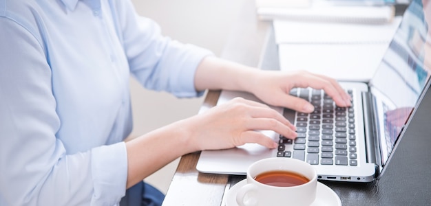 Concepto de negocio. Mujer con camisa azul escribiendo en la computadora con café en la mesa de oficina, retroiluminación, efecto de resplandor del sol, de cerca, vista lateral, espacio de copia