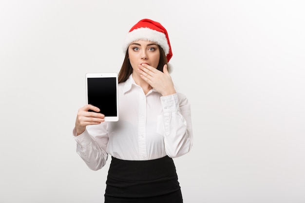 Concepto de negocio - mujer de negocios caucásica joven en tema de Navidad mostrando tableta digital con sorprendente expresión facial.