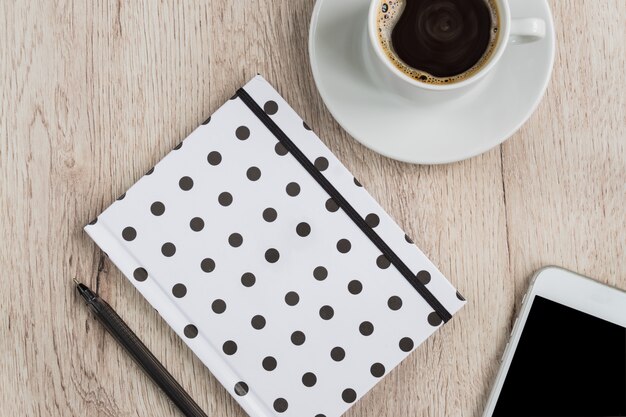 Concepto de negocio y oficina - cuaderno de tapa de lunares en blanco y negro, teléfono inteligente y una taza de café negro en la mesa de madera. vista superior.