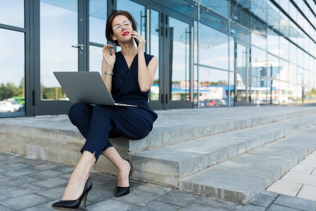 Concepto de negocio. Primer plano de una atractiva joven empresaria con labios rojos con un portátil sentado en los escalones contra la pared de un edificio de oficinas