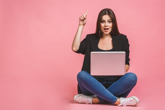 Concepto de negocio. Retrato de mujer feliz en casual sentado en el piso en postura de loto y sosteniendo portátil aislado sobre fondo rosa