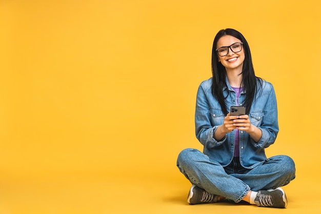 Concepto de negocio Retrato de mujer joven feliz en casual sentado en el suelo en posición de loto y sosteniendo el teléfono móvil aislado sobre fondo amarillo
