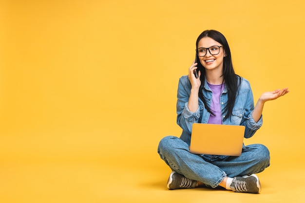 Concepto de negocio Retrato de mujer joven feliz sentada casualmente en el suelo en posición de loto y sosteniendo una computadora portátil aislada sobre fondo amarillo usando teléfono móvil