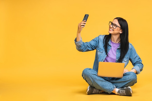 Concepto de negocio Retrato de mujer joven feliz sentada casualmente en el suelo en posición de loto y sosteniendo una computadora portátil aislada sobre fondo amarillo usando teléfono móvil