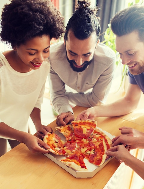 concepto de negocios, comida, almuerzo y personas - equipo de negocios feliz comiendo pizza en la oficina