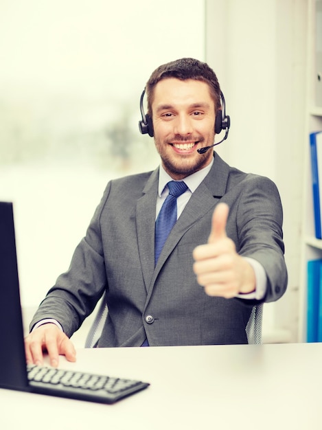 Foto concepto de negocios, comunicación y tecnología: operador de línea de ayuda masculino amigable con auriculares y computadora en el centro de llamadas que muestra el pulgar hacia arriba