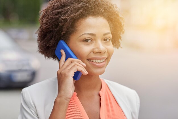 concepto de negocios, comunicación, tecnología y personas - joven mujer de negocios afroamericana sonriente llamando a un teléfono inteligente en la ciudad