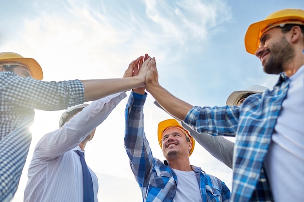 concepto de negocios, construcción, asociación, gestos y personas - cerca de constructores y arquitectos sonrientes en cascos haciendo cinco al aire libre