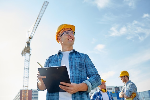 concepto de negocios, construcción, papeleo y personas - constructor feliz en casco con portapapeles y lápiz sobre un grupo de constructores en el sitio de construcción