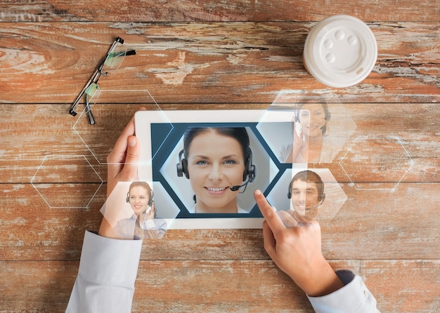 Foto concepto de negocios, personas, comunicación y tecnología - cierre de las manos apuntando con el dedo a la pantalla de la computadora de la tableta con la imagen del operador de la línea de ayuda, taza de café y anteojos en la mesa