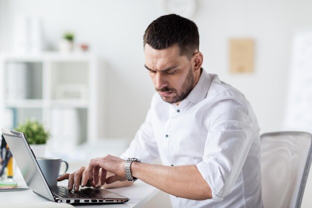 concepto de negocios, personas, fecha límite, tiempo y tecnología: hombre de negocios estresado con computadora portátil mirando su reloj de pulsera en la oficina