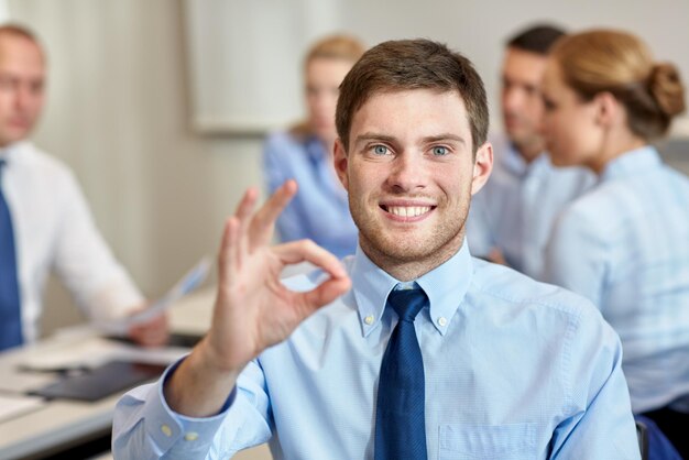 concepto de negocios, personas, gestos y trabajo en equipo - hombre de negocios sonriente que muestra un gesto correcto con un grupo de empresarios reunidos en la oficina