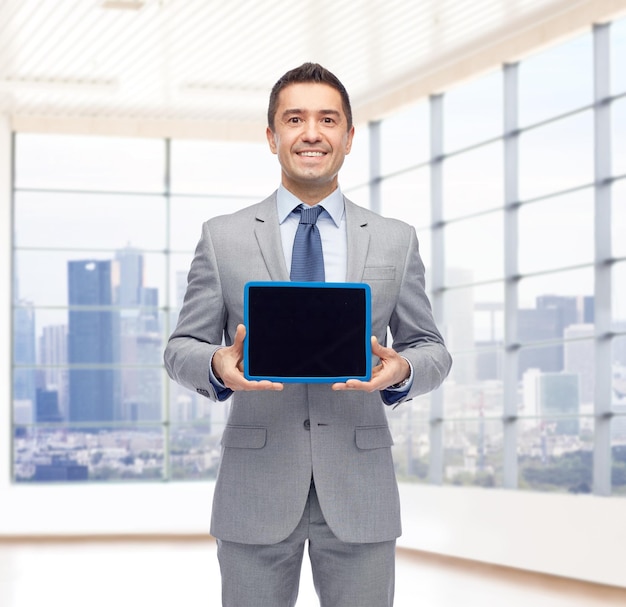 El concepto de negocios, personas y tecnología: feliz hombre de negocios sonriente con traje que muestra una pantalla negra en blanco de una computadora de tablet pc sobre el fondo de la ventana de la oficina de la ciudad