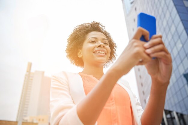 concepto de negocios, tecnología, comunicación y personas - joven mujer de negocios afroamericana sonriente con teléfono inteligente en la ciudad