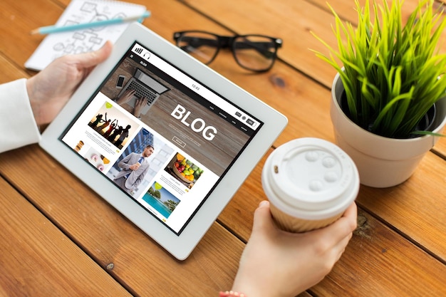 concepto de negocios, tecnología, medios, Internet y personas: cierre de una mujer con una página web de blog en la pantalla de una tableta tomando café en una mesa de madera