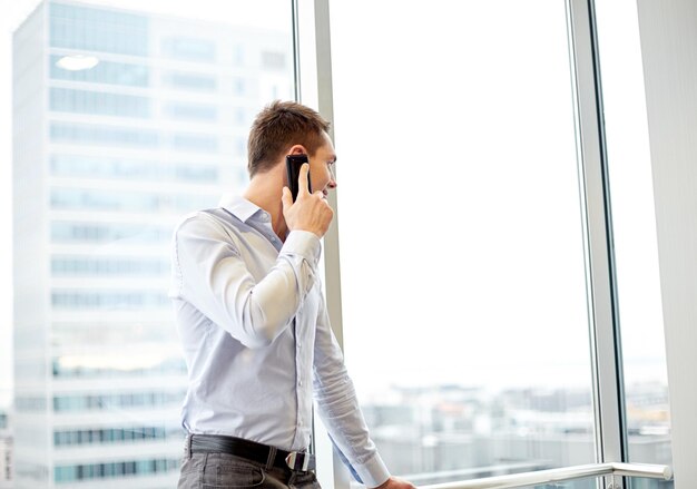 concepto de negocios, tecnología y personas - hombre de negocios feliz llamando a un teléfono inteligente y mirando por la ventana de la oficina