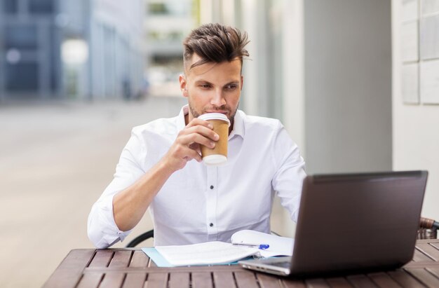 concepto de negocios, tecnología y personas - joven con computadora portátil y taza de café en el café de la calle de la ciudad