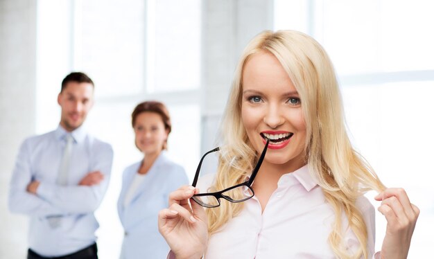 concepto de negocios, trabajo en equipo y personas: mujer de negocios sonriente, estudiante o secretaria con anteojos sobre la oficina y el fondo de un grupo de colegas