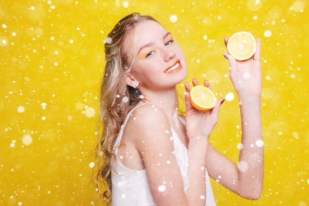 Concepto de nieve, invierno, navidad, emociones, salud, personas, comida y belleza - Beauty Model Girl toma Juicy Oranges. Hermosa chica adolescente alegre con pecas. maquillaje amarillo. sobre fondo de nieve