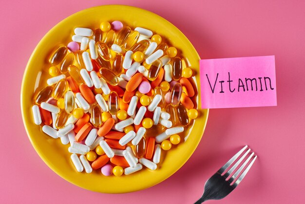 El concepto de nutrición adecuada y medicina. Suplementos dietéticos en cápsulas, pastillas y comprimidos en un plato amarillo sobre fondo rosa. Inscripción Vitaminas en una pegatina.