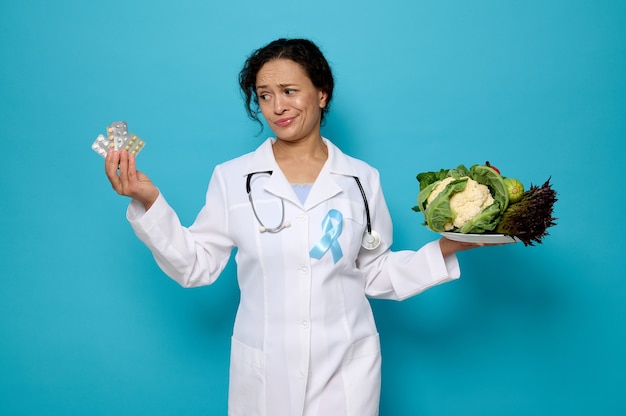 Concepto de nutrición y cuidado de la salud adecuados. Doctora confiada en bata médica con cinta azul de concienciación sobre la diabetes sostiene un plato de comida vegana saludable y una ampolla con medicamentos en sus manos.