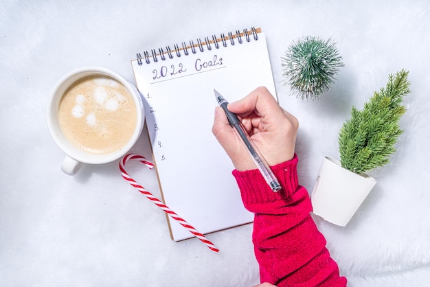 Concepto de objetivos de año nuevo 2022. Plan anual y lista de acciones en el bloc de notas con decoración navideña, taza de café con leche sobre fondo blanco esponjoso flatlay vista superior espacio de copia