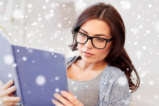 concepto de ocio, educación, visión, invierno y personas - mujer joven con anteojos leyendo un libro en casa sobre la nieve