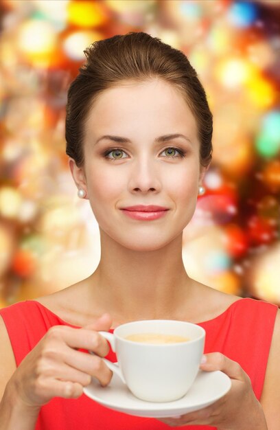 concepto de ocio, felicidad, vacaciones y bebida - mujer sonriente vestida de rojo con una taza de café sobre fondo de luces brillantes