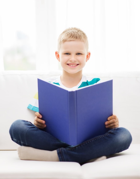 concepto de ocio, infancia, educación y hogar - niño pequeño sonriente leyendo un libro y sentado en el sofá en casa