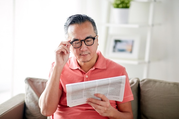 Concepto de ocio, información, personas y medios de comunicación: hombre feliz con gafas leyendo el periódico en casa