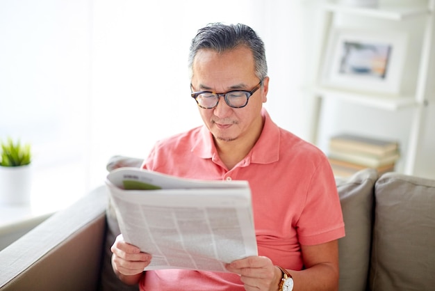Concepto de ocio, información, personas y medios de comunicación: hombre feliz con gafas leyendo el periódico en casa