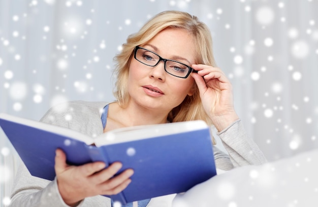 concepto de ocio, literatura y gente - mujer de mediana edad leyendo un libro y sentada en el sofá en casa sobre la nieve