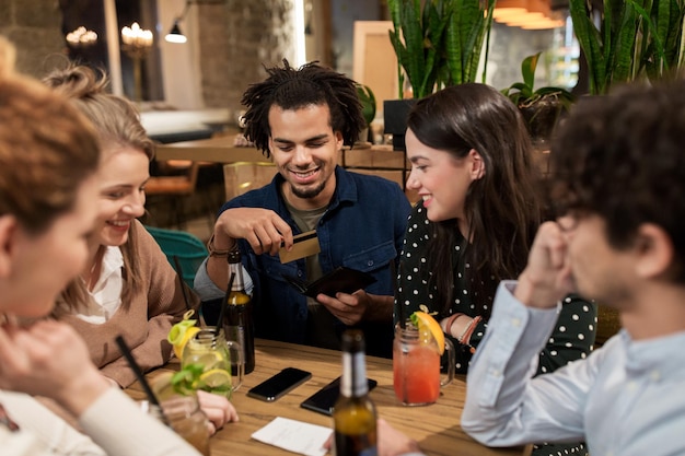 concepto de ocio, personas y vacaciones - amigos felices con bebidas, tarjeta de crédito y factura en un bar o cafetería