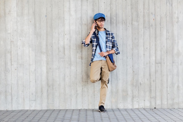concepto de ocio, tecnología, comunicación y personas - hombre hipster sonriente con bolso de hombro llamando al teléfono inteligente en la pared de la calle